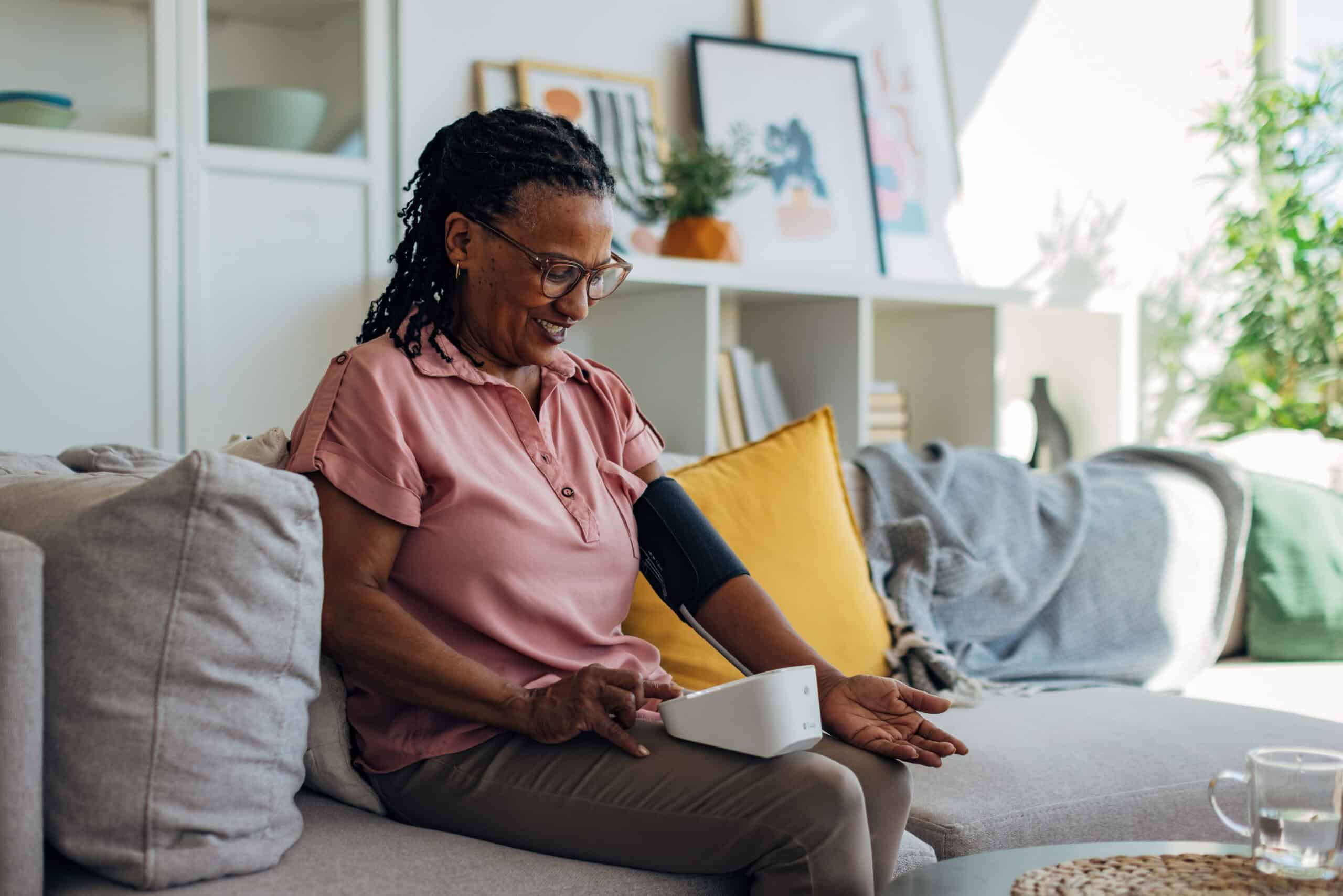 Black woman sits in a peaceful home environment, checking her blood pressure and pulse using a modern digital monitor. The device is secured around her arm, and the display shows both systolic and diastolic pressure along with heart rate